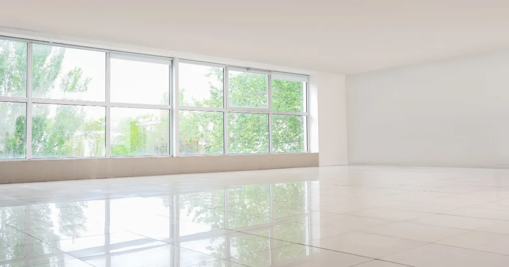 Empty room with white walls, glossy light colored floor with floor to ceiling windows that show how to use soft neutral colors and plenty of natural light to make a room look bigger. 